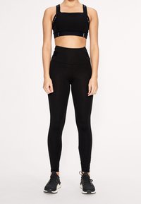 Černá sportovní podprsenka s širokými ramínky a akcenty na sponách, spárovaná s černými legínami s vysokým pasem. Obě mají texturovaný vzhled.