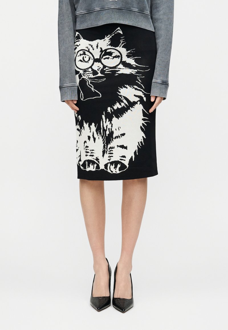 Μαύρη φούστα έως το γόνατο με μεγάλο λευκό γατί που φορά στρογγυλά γυαλιά τυπωμένο στο μπροστινό μέρος, συνδυασμένη με γκρι μακρυμάνικη μπλούζα και μαύρες γόβες.