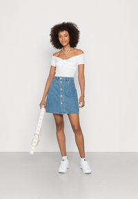 Top blanco de off-the-shoulder con detalle de nudo, combinado con una falda de mezclilla azul con botones frontales. La modelo sostiene un bolso de cintura blanco y lleva zapatillas blancas.