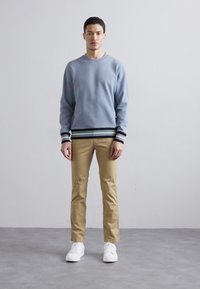 Light blue sweatshirt with black and white striped cuffs, paired with khaki chinos and white sneakers. Model stands against a neutral background.