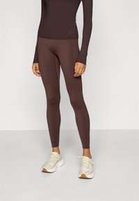 Braune Leggings aus einem dehnbaren Material, mit einem nahtlosen Design und hohem Bund. Getragen mit hellgrauen Sportschuhen.
