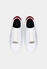 Sneaker bianche con tomaia in pelle liscia, lacci beige e un accento rosso sul tallone. Presentano una placca con logo dorato di lato e suola testurizzata.