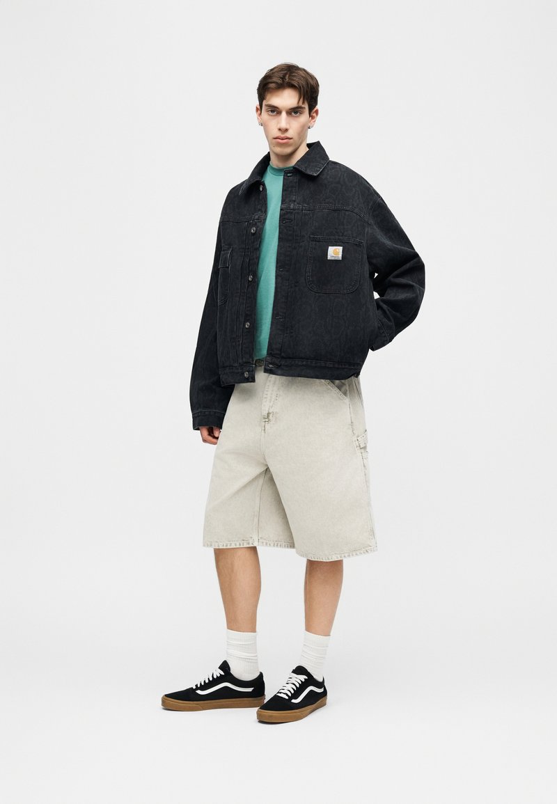 Jonge man die een zwarte spijkerjasje draagt, een teal shirt, beige knielange shorts, witte sokken en zwarte sneakers, staand met één hand in de zak.