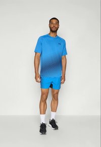 Kék sportpóló, degradált csíkos mintával, kiegészítve hasonló kék rövidnadrággal, fekete részletekkel. A modellen fekete sportcipő van.