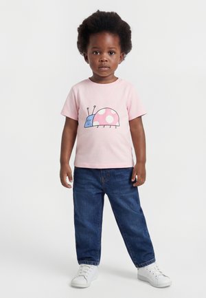 Enfant aux cheveux bouclés et foncés, debout, portant un t-shirt rose avec un insecte coloré de dessin animé, un jean bleu et des baskets blanches sur un fond blanc.