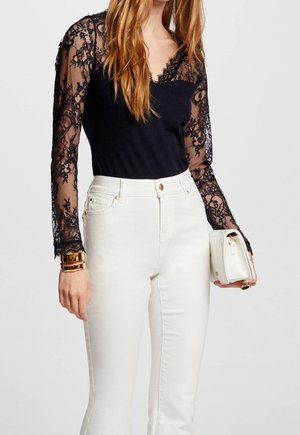 Woman wearing black lace long-sleeve top with scalloped neckline and white high-waist pants, holding a small white handbag and gold bracelets.