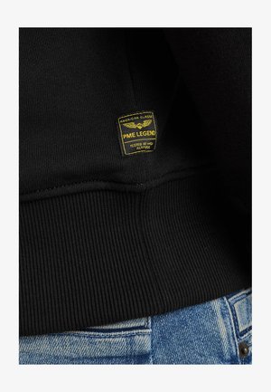 Musta huppari, jossa on ribbireunus ja teksturoitu kangas. Keltainen PME Legend -tunnus, jossa on siivet ja teksti "Kokeiltu korkealla korkeudella".