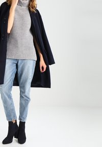 Grå ribbade turtleneck-tröja kombinerad med ljusblå slitna jeans och en marinblå kappa. Svarta ankelskor fulländar outfiten.