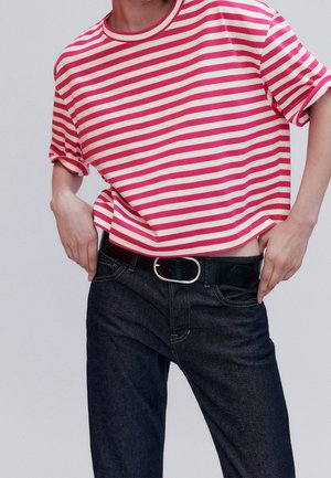 Personne portant un t-shirt rayé rouge et blanc rentré dans un jean foncé avec une ceinture noire, mains ajustant la taille.