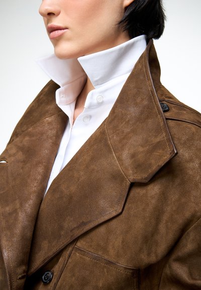 Közeli kép egy személyről, aki fehér galléros inget visel barna szarvasbőr dzseki alatt, széles hajtókákkal, bemutatva a rétegzett divat részleteit.