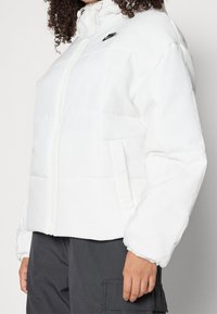 Vit pufferjacka med hög krage, dragkedja framtill och sidofickor. Har en svart Nike-logotyp på övre vänstra bröstet.