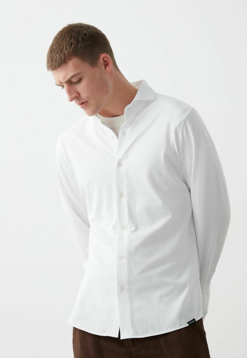 Witte lange mouwen shirt met een button-down kraag, gemaakt van gladde stof, met een subtiele glans en standaard knoopsluiting. Donkerbruine broek daaronder.