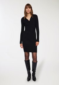 Abito knit nero a coste con scollo a V, maniche lunghe e silhouette aderente, abbinato a stivali neri al ginocchio. Texture liscia e colore uniforme.