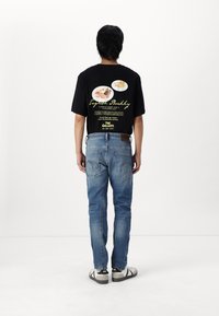 Schwarzes Grafik-T-Shirt mit Lebensmittelillustrationen und Text auf dem Rücken. Tragen mit ausgewaschenen blauen Jeans und weißen Sneakers mit schwarzen Akzenten.