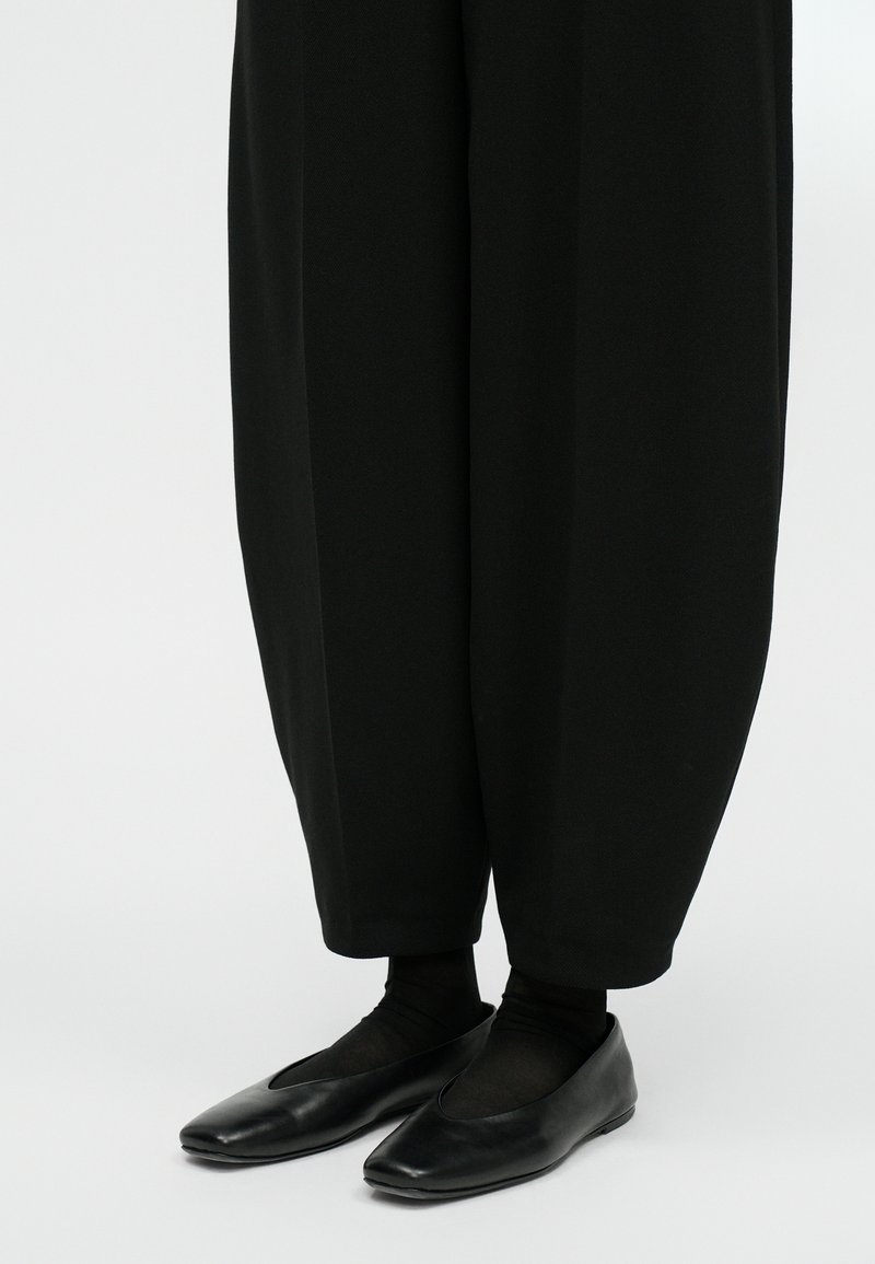 Pantaloni neri a gamba larga con una finitura testurizzata, abbinati a mocassini neri con punta affusolata e superficie in pelle liscia.