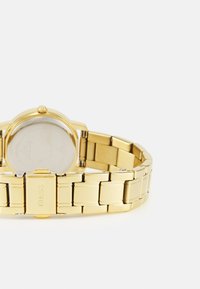 Gold bracelet watch with a round face, polished metal links, and a fold-over clasp. Features a clear watch face and branding on the back.