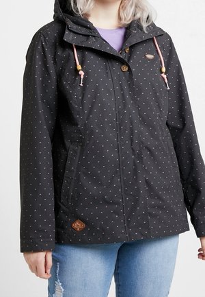 Kobieta w czarnej kurtce z kapturem z małymi białymi wzorami, różowymi sznureczkami i jasno-niebieskich podartych dżinsach, stojąca na białym tle.