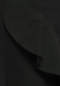 Zwarte blazer van een gestructureerde, gestreepte stof. Heeft een grote, vloeiende ruches aan één kant. Gladde textuur met een subtiele glans.