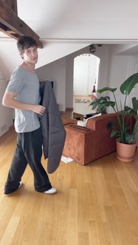 Grå vadderad jacka hålls av en person, kombinerad med svarta byxor och vita skor. Rummet inkluderar en manchestersoffa och en stor grön växt.