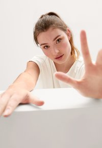 Witte korte mouwen top, model leunend naar voren met handen op een witte oppervlak. Zachte huidtextuur, neutrale achtergrond. Close-up perspectief.