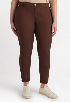 Pantalones marrones ajustados, hechos de una tela suave, con cremallera, cierre de botón y bolsillos laterales. Combinados con zapatillas blancas.