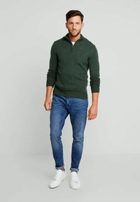 Green knit sweater with a half-zip collar, paired with blue slim-fit jeans and white sneakers; smooth texture, fitted design.