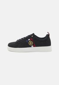 Sneaker noir avec une tige texturée, présente un emblème doré, un accent rayé en bleu, blanc et rouge sur le côté, et une semelle extérieure blanche.