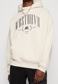 Felpa crema realizzata in tessuto morbido, con tasca frontale, design oversize e stampa del marchio in grigio scuro. Accentuata da un cappuccio con cordino.