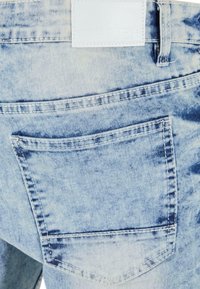 Jean en denim bleu clair avec une seule poche arrière. La texture du tissu est légèrement usée avec des coutures visibles et une étiquette blanche.