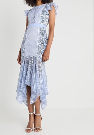 Frau trägt ein hellblaues, florales Spitzenkleid mit flatternden Ärmeln, tailliertem Schnitt und asymmetrischem Saum, kombiniert mit silbernen Knöchelriemen-Pumps.