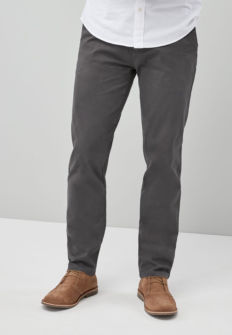 Graue Baumwollhose mit Slim-Fit, glatter Textur und Fronttaschen, kombiniert mit braunen Wildlederschuhen. Der Bund verfügt über Gürtelschlaufen.