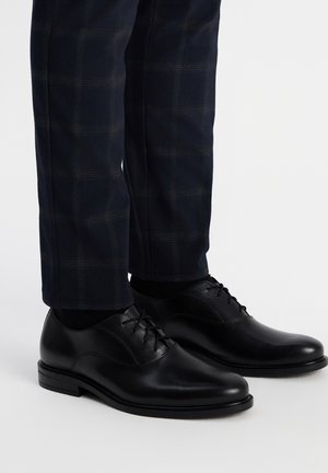 Czarne skórzane buty wizytowe o gładkiej fakturze, okrągłych czubkach i sznurówkach. Noszone z ciemnymi spodniami w kratę, z cienkimi liniami jaśniejszego szarości.