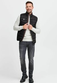 Schwarze gepolsterte Weste mit hohem Kragen, Reißverschluss vorne und Taschen an den Seiten. Getragen über einem hellgrauen Pullover und kombiniert mit dunkelgrauen Jeans und schwarzen Schuhen.