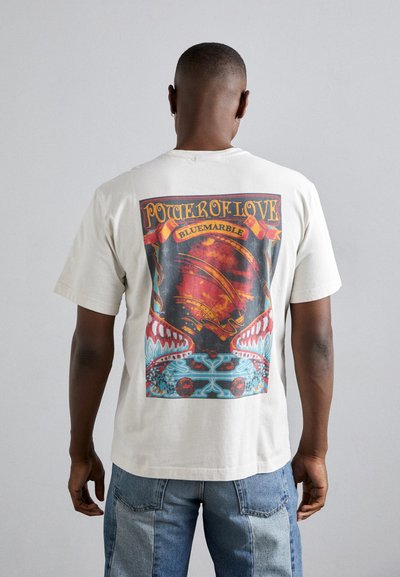 Cremefarvet t-shirt med et stort grafisk print på ryggen, med klare farver og detaljerede design, herunder røde, blå og gyldne accenter.