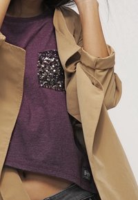 Lila T-Shirt mit einer Pailletten-Tasche, darunter eine sandfarbene Trenchjacke mit hochgekrempelten Ärmeln. Weiche Stoffstruktur; lässige Passform.