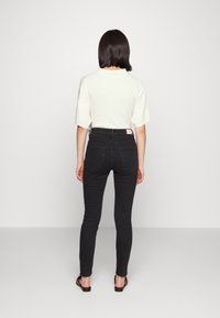 Kvinna som står vänd mot en vit vägg, iklädd en vit kort-ärmad topp, svarta högmidjade skinny jeans och svarta platta sandaler.