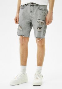 Mies, joka on pukeutunut vaaleanharmaisiin revittyihin farkkushortseihin, valkoisiin sukkiin ja valkoisiin tennareihin, seisoo pelkää valkoista taustaa vasten.