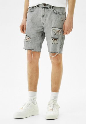 Man som bär ljusgrå slitna denimshorts, vita strumpor och vita sneakers, stående mot en enfärgad vit bakgrund.