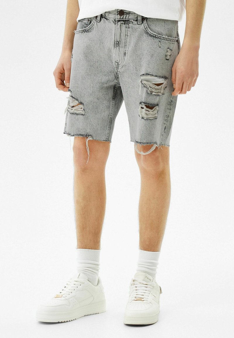 Mies, joka on pukeutunut vaaleanharmaisiin revittyihin farkkushortseihin, valkoisiin sukkiin ja valkoisiin tennareihin, seisoo pelkää valkoista taustaa vasten.