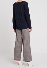 Henkilö, jolla on päällään tummansininen pitkähihaisen paita, leveälahkeiset ruudulliset housut ja valkoiset korkeat nilkkabootsit, seisoo yksinkertaista taustaa vasten.