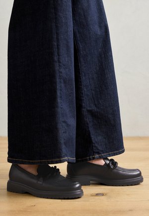 Mocassins en cuir noir avec une finition texturée et un nœud décoratif. Semelle en caoutchouc épaisse. Associés à un pantalon en denim large et foncé.