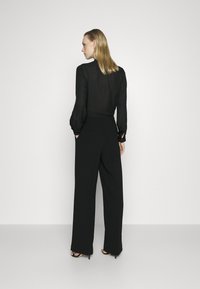 Blouse noire transparente avec des manches bouffantes, associée à un pantalon noir taille haute. Le modèle se tient de profil devant un fond blanc uni.
