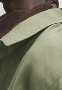 Personne portant une écharpe marron foncé et un manteau vert clair avec un col visible, vue en contre-plongée en gros plan.