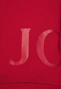 Tissu rouge avec de grandes lettres brillantes "J" et une partie de "O" imprimées sur la surface, montrant la texture et un léger éclat.
