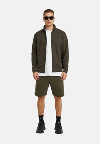 Giacca zip-up verde oliva sopra una maglietta bianca, abbinata a pantaloni corti coordinati. Occhiali da sole neri e scarpe da ginnastica completano l'outfit.