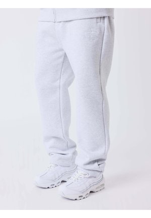 Jambes inférieures portant un pantalon de survêtement gris clair avec un petit logo et des baskets blanches sur un fond uni.
