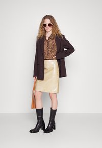 Rjav oversized blazer, srajca z živalskim vzorcem, sijoča bež usnjena krilo, črne škornje na peto in velike bele sončne očala. Rjav nahrbtnik v roki.