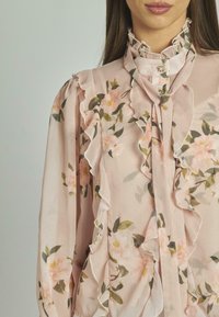 Blusa floreale rosa in chiffon, con volant lungo il fronte e il collo, dettagli a bottoni e una texture morbida e leggera.