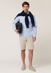 Heren lichtblauw overhemd met knopen, beige shorts, marineblauwe gebreide trui over de schouders, zwarte tas en lichtgrijze instappers.