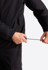 Άτομο που σφίγγει το ελαστικό κορδόνι της μανσέτας ενός μαύρου μπουφάν σε απλό φωτεινό φόντο, φορώντας ταιριαστό μαύρο παντελόνι με τσέπη.